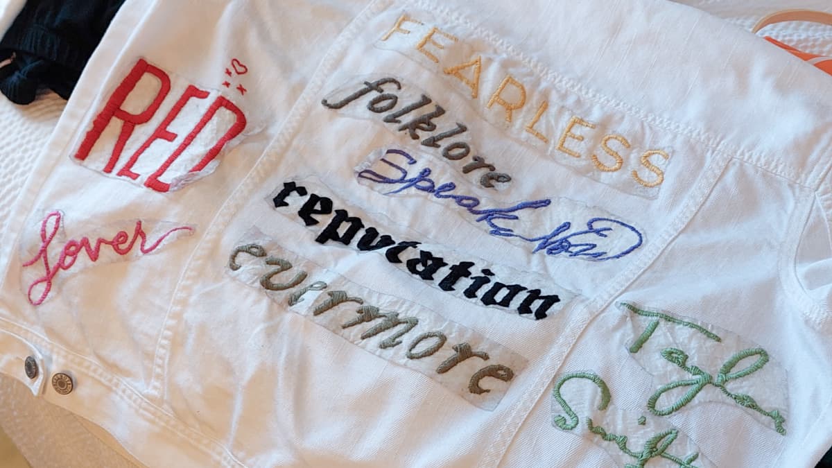Close-up picture of white jean jacket with Taylor Swift album titles embroidered on it.