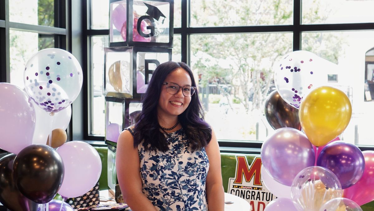 Yasmin is smiling at the camera, with purple, pink, and gold balloons behind her.